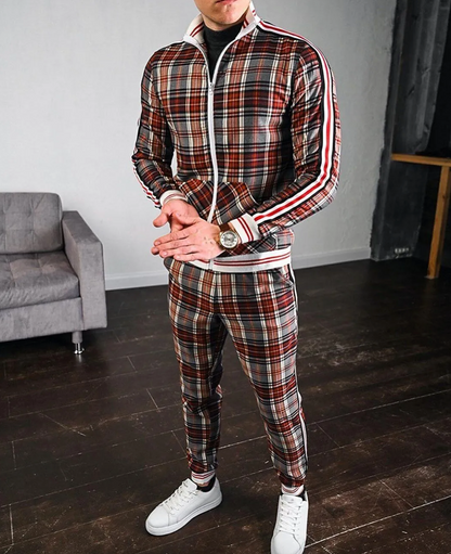 Uomo in tuta a quadri rossi e bianchi, con orologio al polso, in piedi su pavimento in legno, vicino a divano grigio. Moda casual maschile.