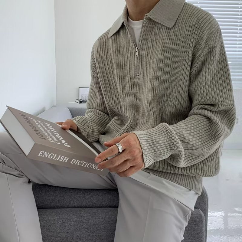 Uomo seduto con maglione grigio a maglia, pantaloni chiari, tiene un dizionario inglese. Interno moderno, luce naturale.