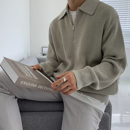 Uomo seduto con maglione grigio a maglia, pantaloni chiari, tiene un dizionario inglese. Interno moderno, luce naturale.