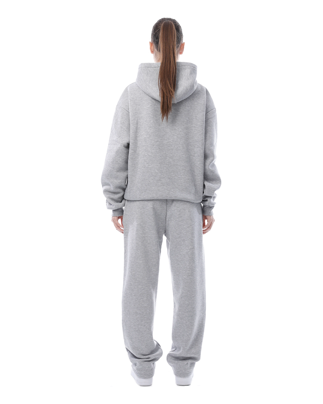 Donna in tuta grigia, vista posteriore. Abbigliamento sportivo casual, felpa con cappuccio e pantaloni larghi. Stile comodo e moderno.