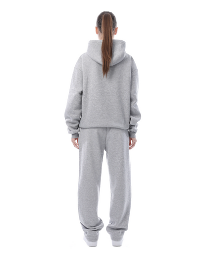 Donna in tuta grigia, vista posteriore. Abbigliamento sportivo casual, felpa con cappuccio e pantaloni larghi. Stile comodo e moderno.