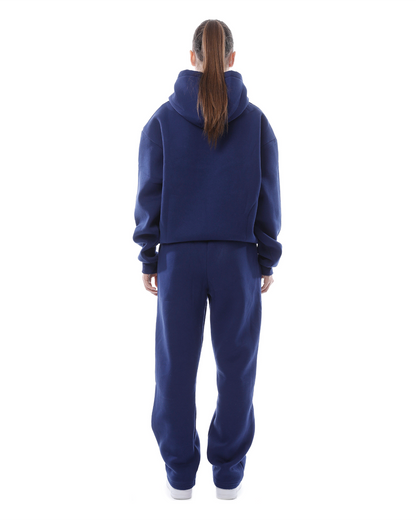 Donna di spalle con felpa e pantaloni blu, capelli raccolti, su sfondo bianco. Abbigliamento casual, moda autunnale, stile sportivo.