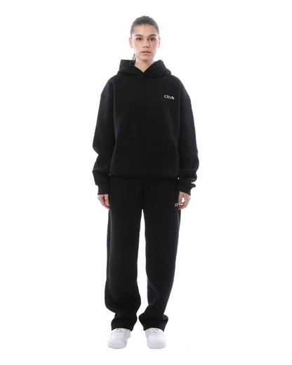 Donna in tuta nera con cappuccio, pantaloni larghi e scarpe bianche. Abbigliamento casual, moda streetwear, sfondo bianco.