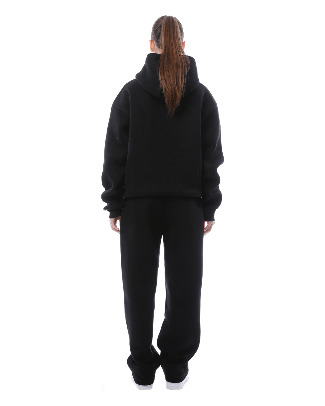 Donna di spalle con felpa nera con cappuccio e pantaloni abbinati, su sfondo bianco. Abbigliamento casual, moda streetwear, look total black.