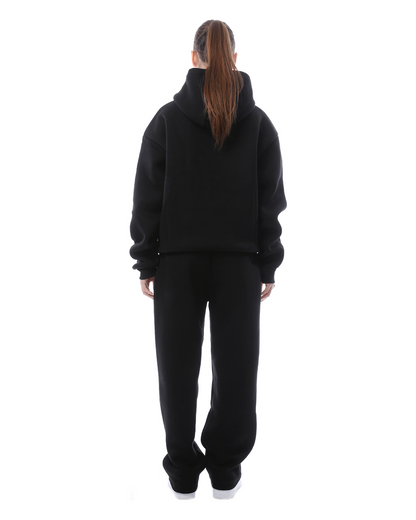Donna di spalle con felpa nera con cappuccio e pantaloni abbinati, su sfondo bianco. Abbigliamento casual, moda streetwear, look total black.