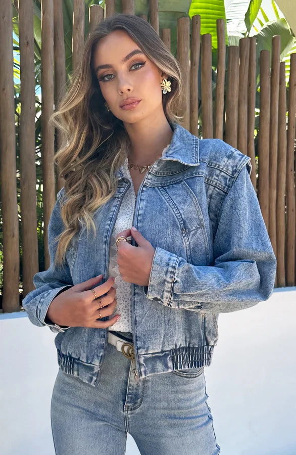 Donna con giacca di jeans, camicia bianca e jeans, posa all'aperto davanti a una staccionata di legno. Moda casual, stile denim, look estivo.
