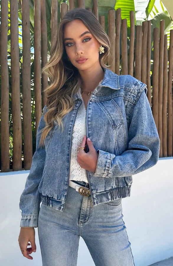 Donna con giacca di jeans, camicia bianca e jeans, posa all'aperto. Moda casual, stile denim, look primaverile. Sfondo con piante e recinzione in legno.