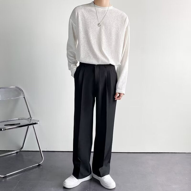 Uomo in abbigliamento casual elegante, indossa maglia bianca e pantaloni neri larghi, con collana argento. Sfondo minimalista, sedia pieghevole.
