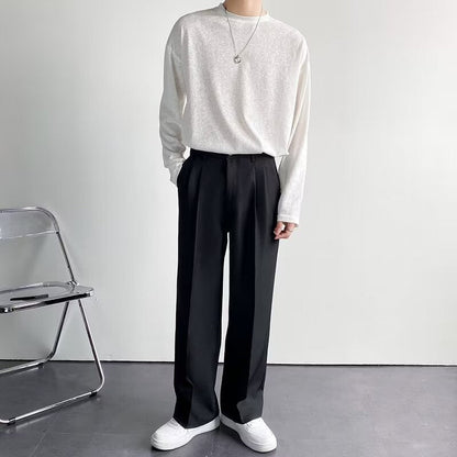Uomo in abbigliamento casual elegante, indossa maglia bianca e pantaloni neri larghi, con collana argento. Sfondo minimalista, sedia pieghevole.