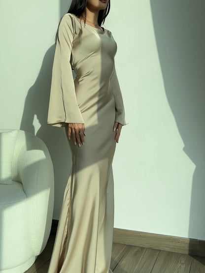 Donna in abito lungo beige elegante, maniche svasate, in piedi accanto a poltrona bianca. Moda femminile, abbigliamento da sera, stile sofisticato.