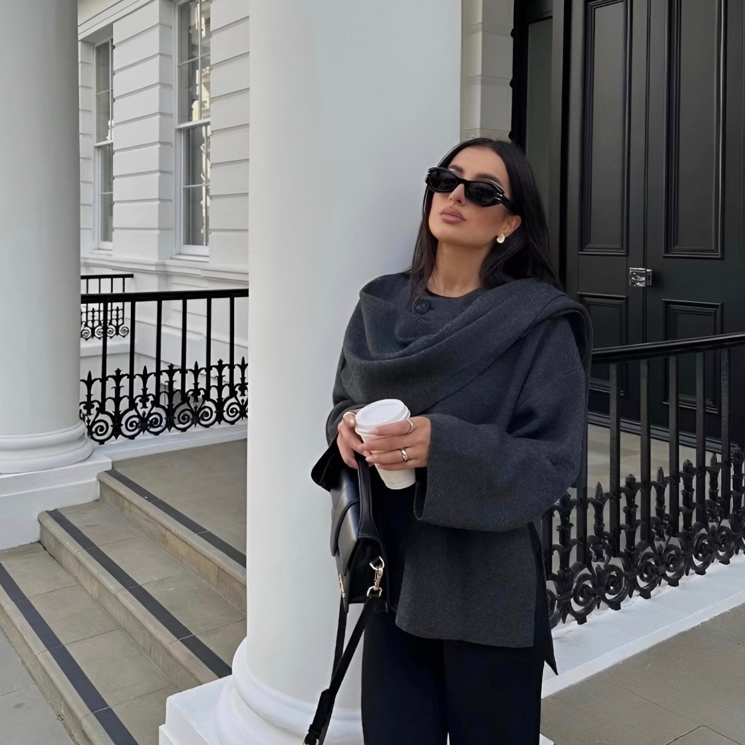 Donna elegante con occhiali da sole, cappotto grigio e borsa nera, tiene una tazza di caffè davanti a un edificio classico bianco con colonne.