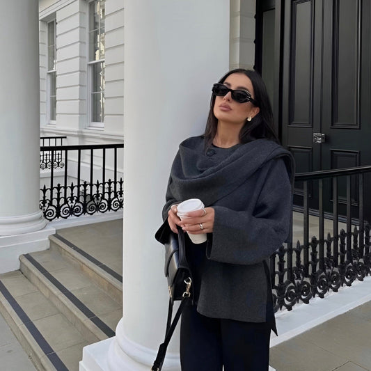 Donna elegante con occhiali da sole, cappotto grigio e borsa nera, tiene una tazza di caffè davanti a un edificio classico bianco con colonne.