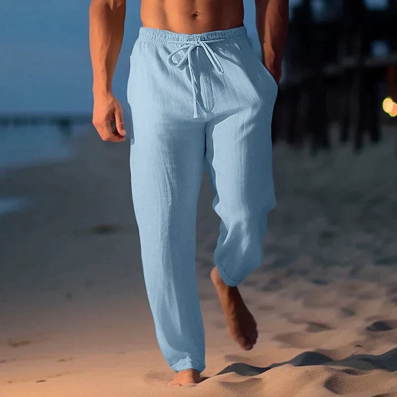Uomo che cammina sulla spiaggia al tramonto indossando pantaloni di lino azzurri, moda estiva maschile, abbigliamento casual elegante.
