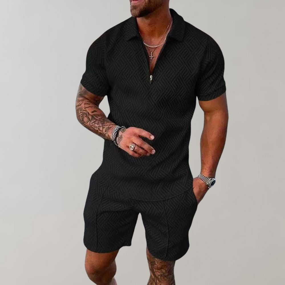 Uomo muscoloso in tuta nera elegante con motivo geometrico, braccia tatuate, collane e orologio, posa su sfondo grigio chiaro.