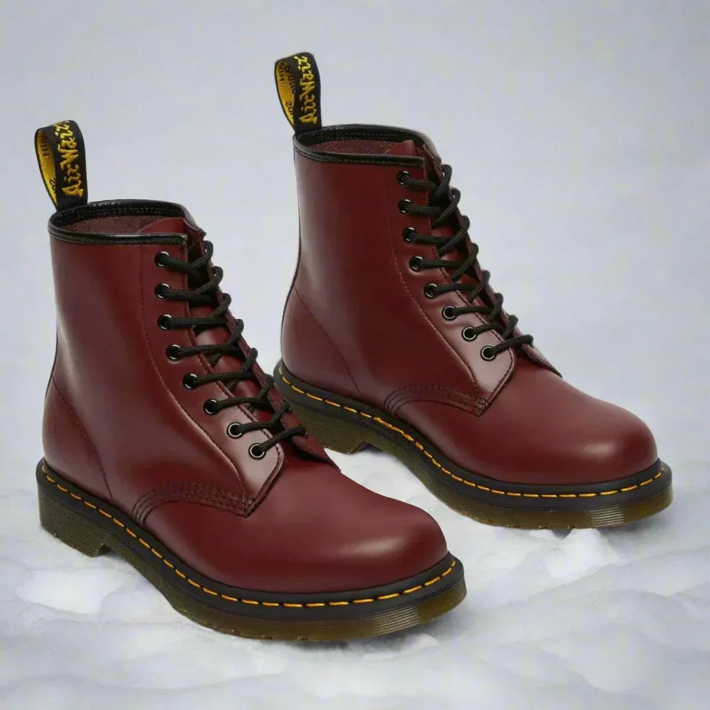 Stivali Dr. Martens rossi in pelle, suola in gomma nera con cuciture gialle, lacci neri, etichetta posteriore gialla, su sfondo bianco.