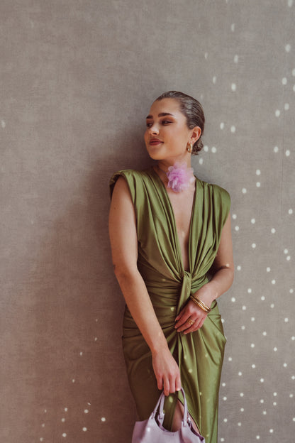 Donna in abito verde elegante con scollo profondo, accessori oro e borsa lilla, posa contro parete grigia con luci puntiformi. Moda sofisticata.