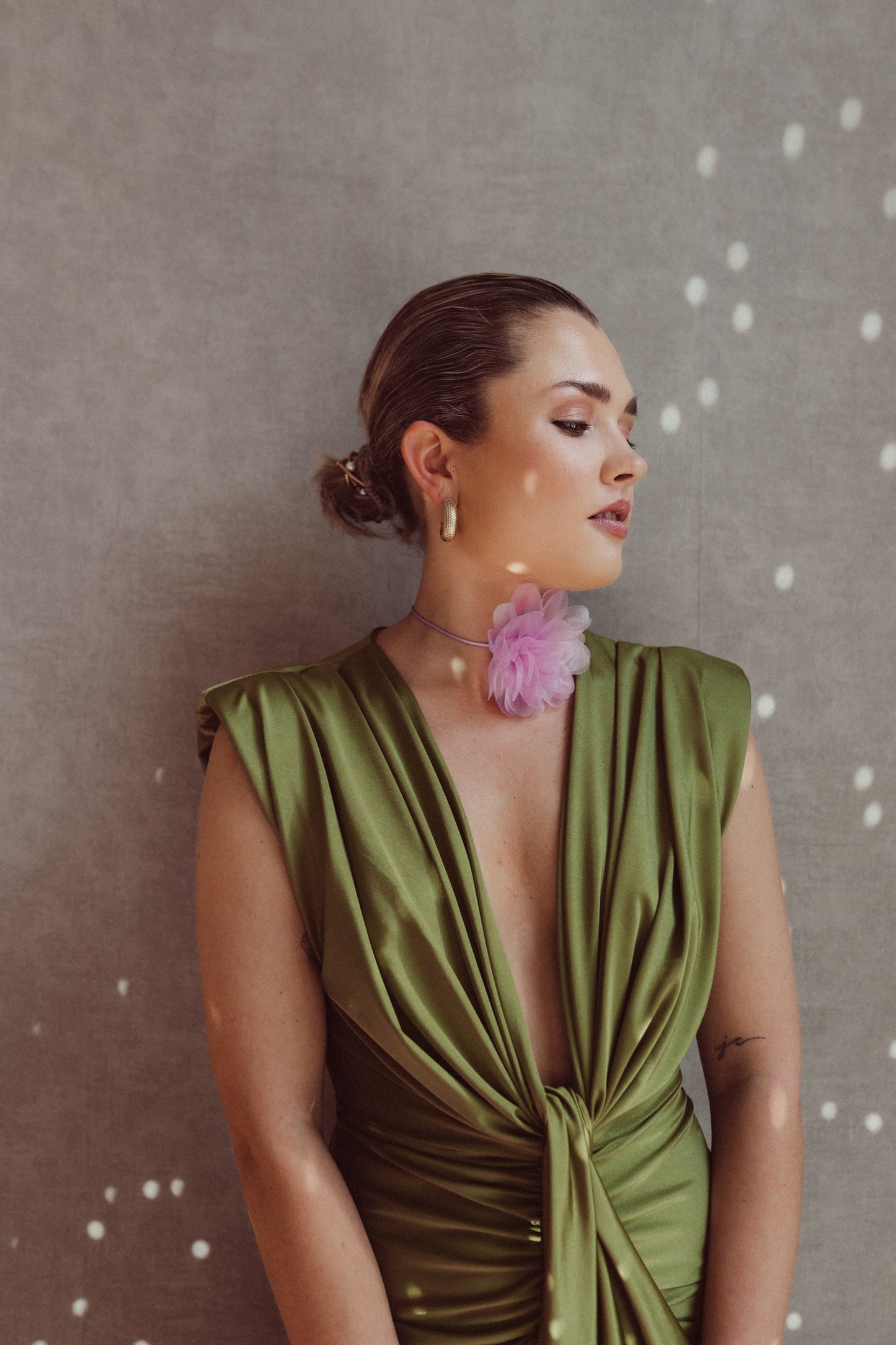 Donna elegante in abito verde satinato con scollatura profonda, indossa orecchini dorati e collana con fiore rosa, sfondo grigio con luci soffuse.