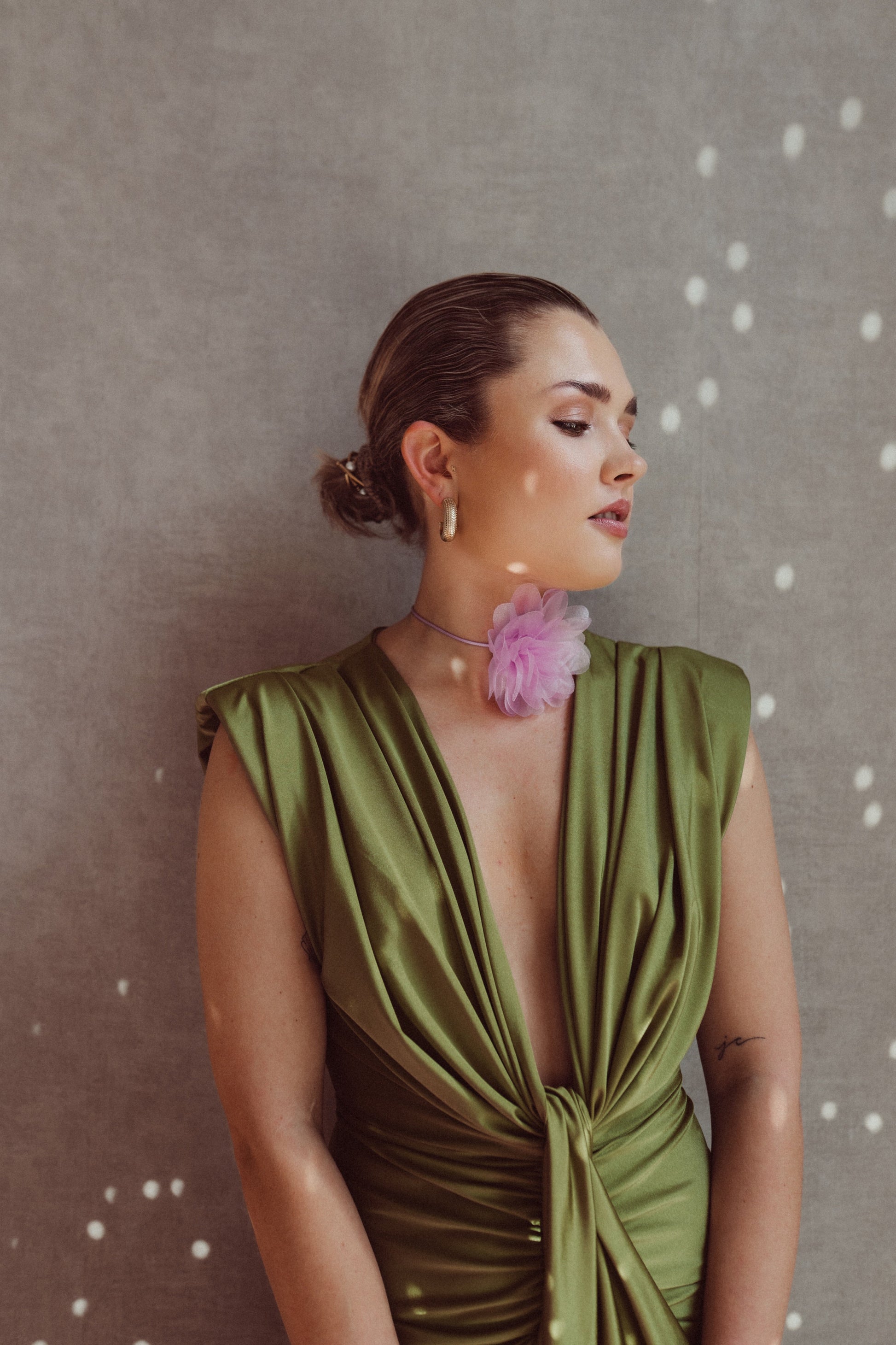 Donna elegante in abito verde satinato con scollatura profonda, indossa orecchini dorati e collana con fiore rosa, sfondo grigio con luci soffuse.