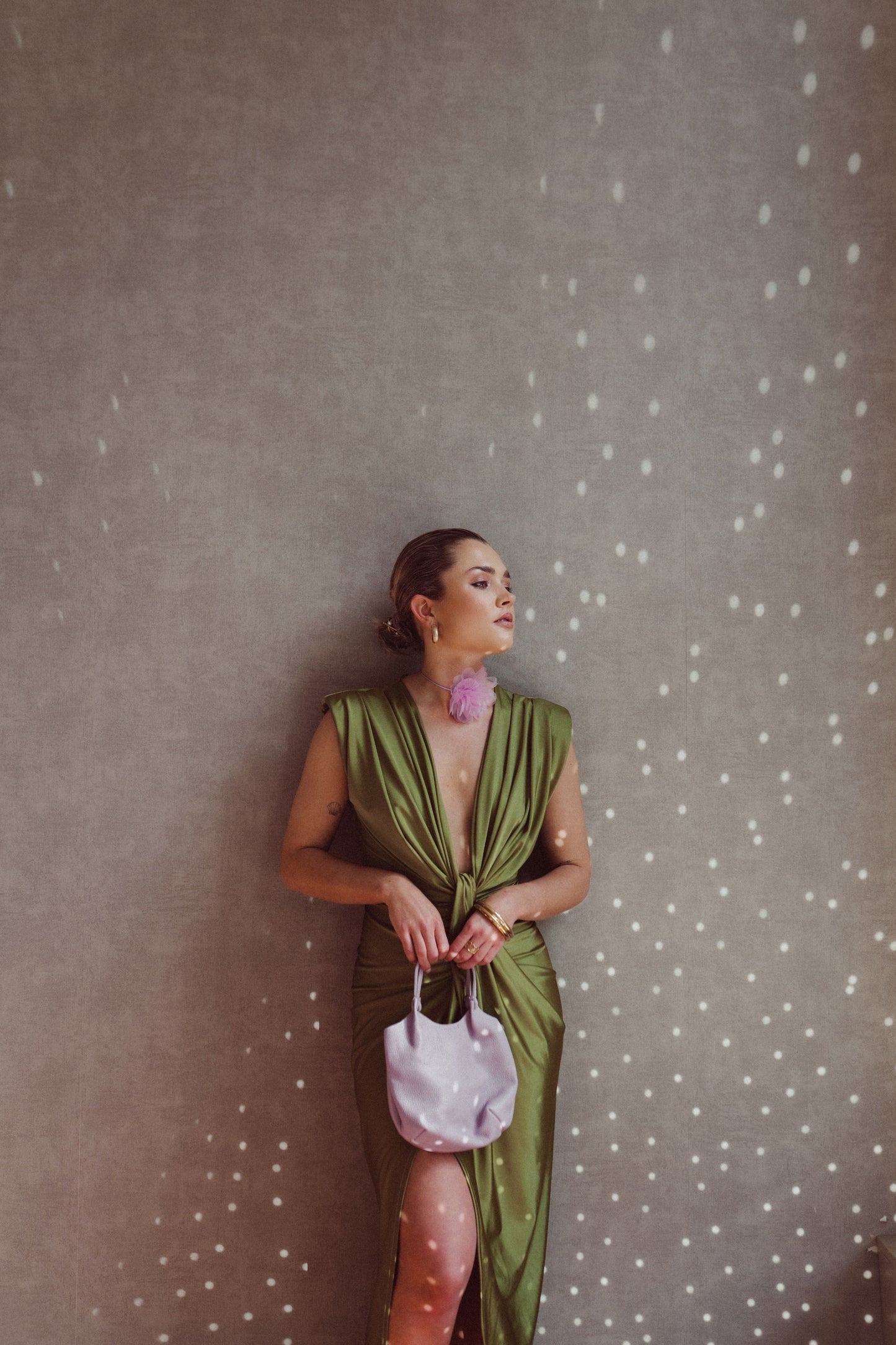 Donna elegante in abito verde satinato con spacco, borsa bianca e orecchini rosa, appoggiata a parete con luce puntinata. Moda sofisticata.