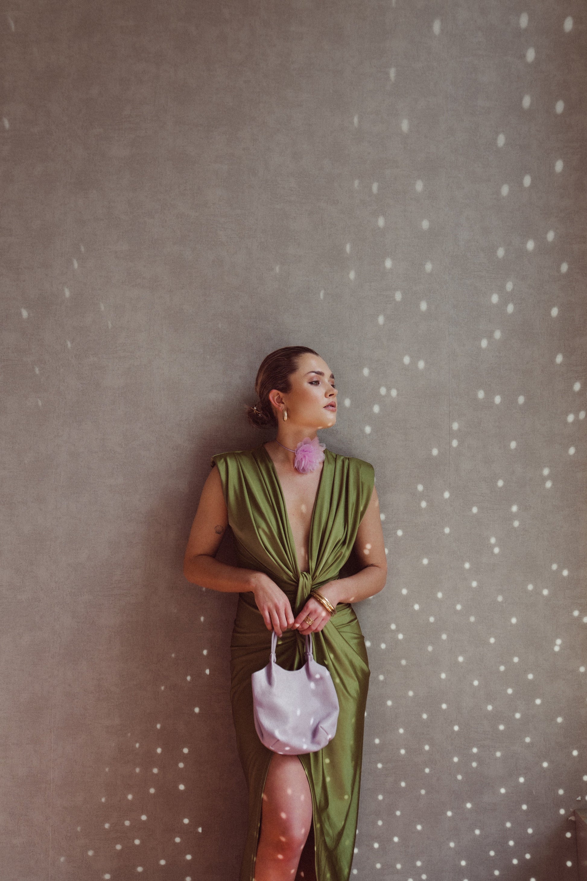 Donna elegante in abito verde satinato con spacco, borsa bianca e orecchini rosa, appoggiata a parete con luce puntinata. Moda sofisticata.