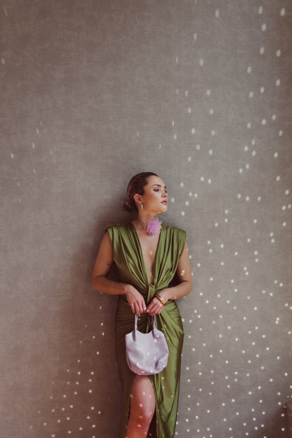 Donna elegante in abito verde satinato con spacco, borsa bianca e orecchini rosa, appoggiata a parete con luce puntinata. Moda sofisticata.