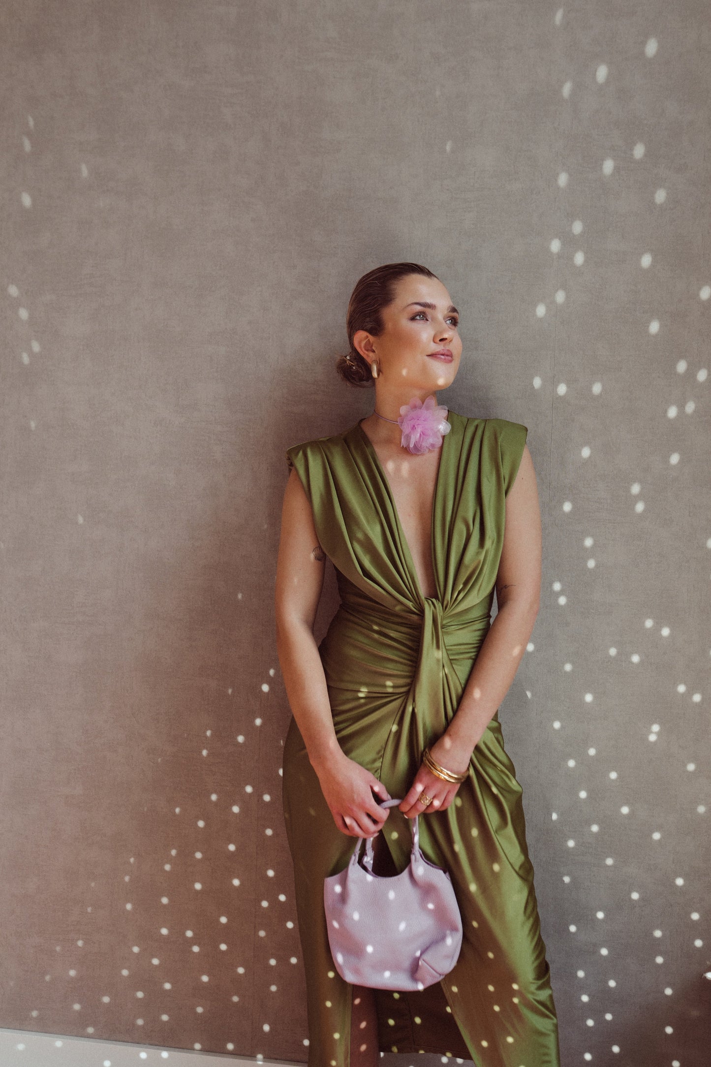 Donna elegante in abito verde drappeggiato, accessoriata con borsa rosa e orecchino floreale, posa contro parete con effetto luce puntinato.