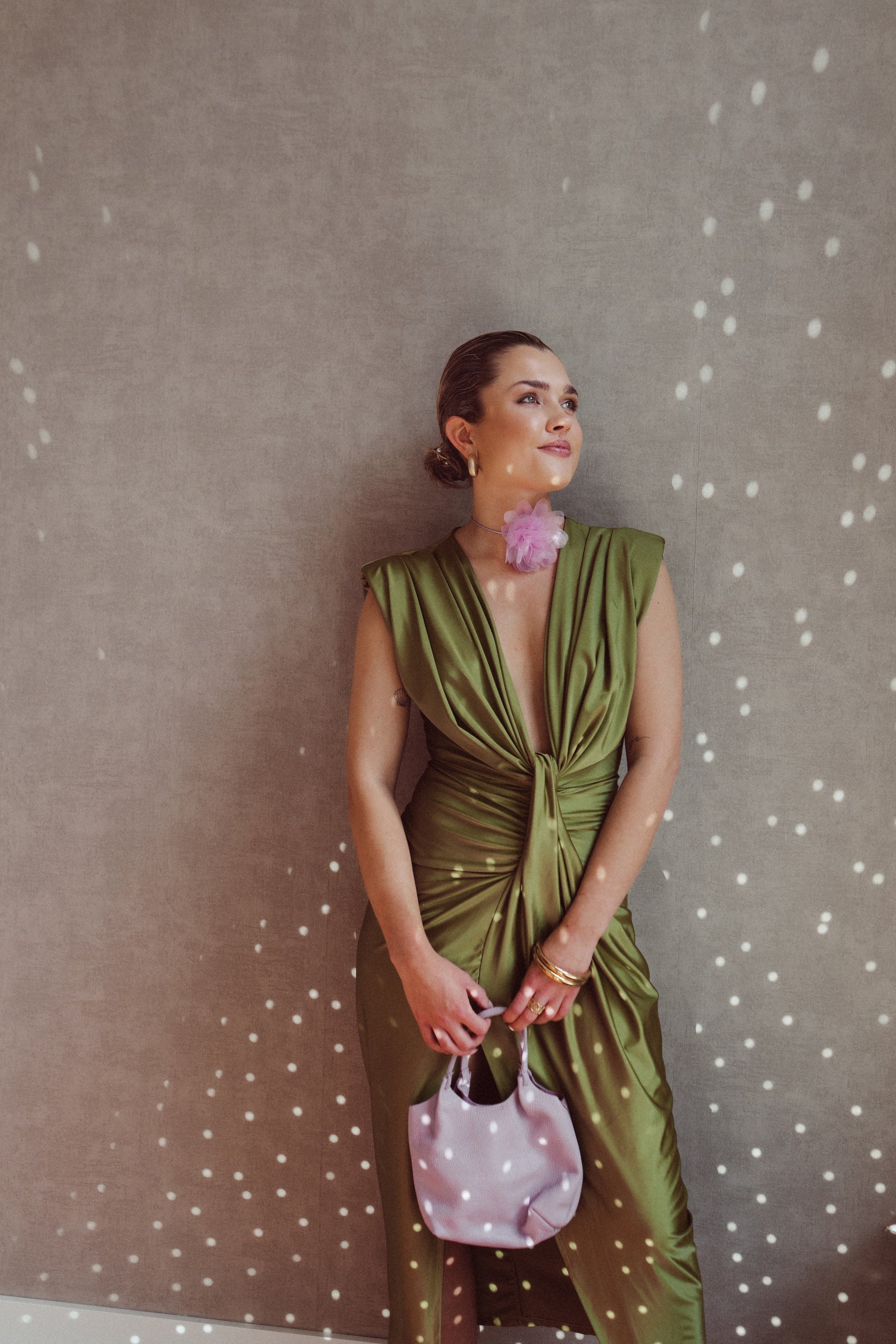 Donna elegante in abito verde drappeggiato, accessoriata con borsa rosa e orecchino floreale, posa contro parete con effetto luce puntinato.