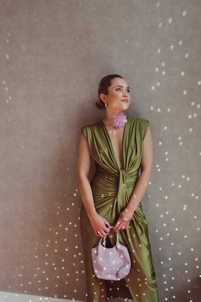 Donna elegante in abito verde drappeggiato, accessoriata con borsa rosa e orecchino floreale, posa contro parete con effetto luce puntinato.