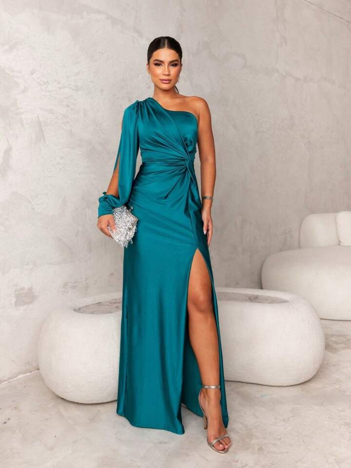 Donna in elegante abito lungo verde smeraldo con spacco, sandali argento e pochette brillante, posa in interni moderni con pareti grigie.