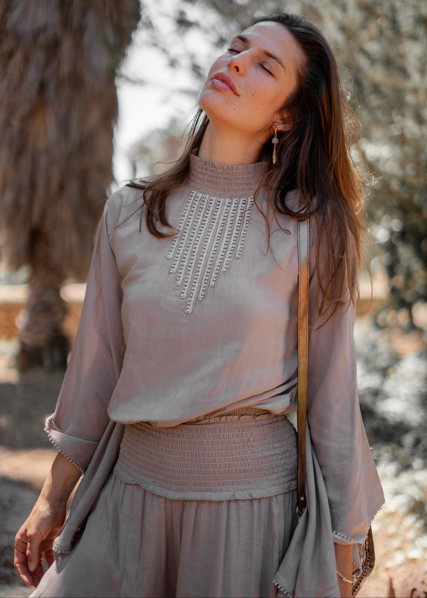 Donna con abito beige elegante, dettagli ricamati, in posa serena all'aperto. Moda femminile, stile boho-chic, natura sullo sfondo.