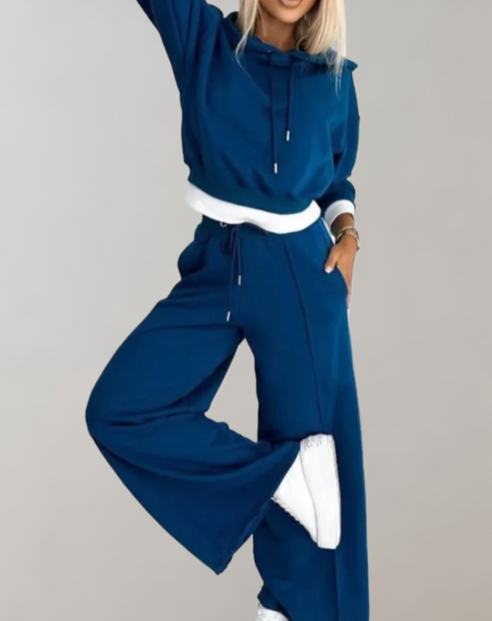 Donna in tuta sportiva blu, felpa con cappuccio e pantaloni larghi, posa su sfondo grigio. Moda casual, abbigliamento comodo, stile urbano.