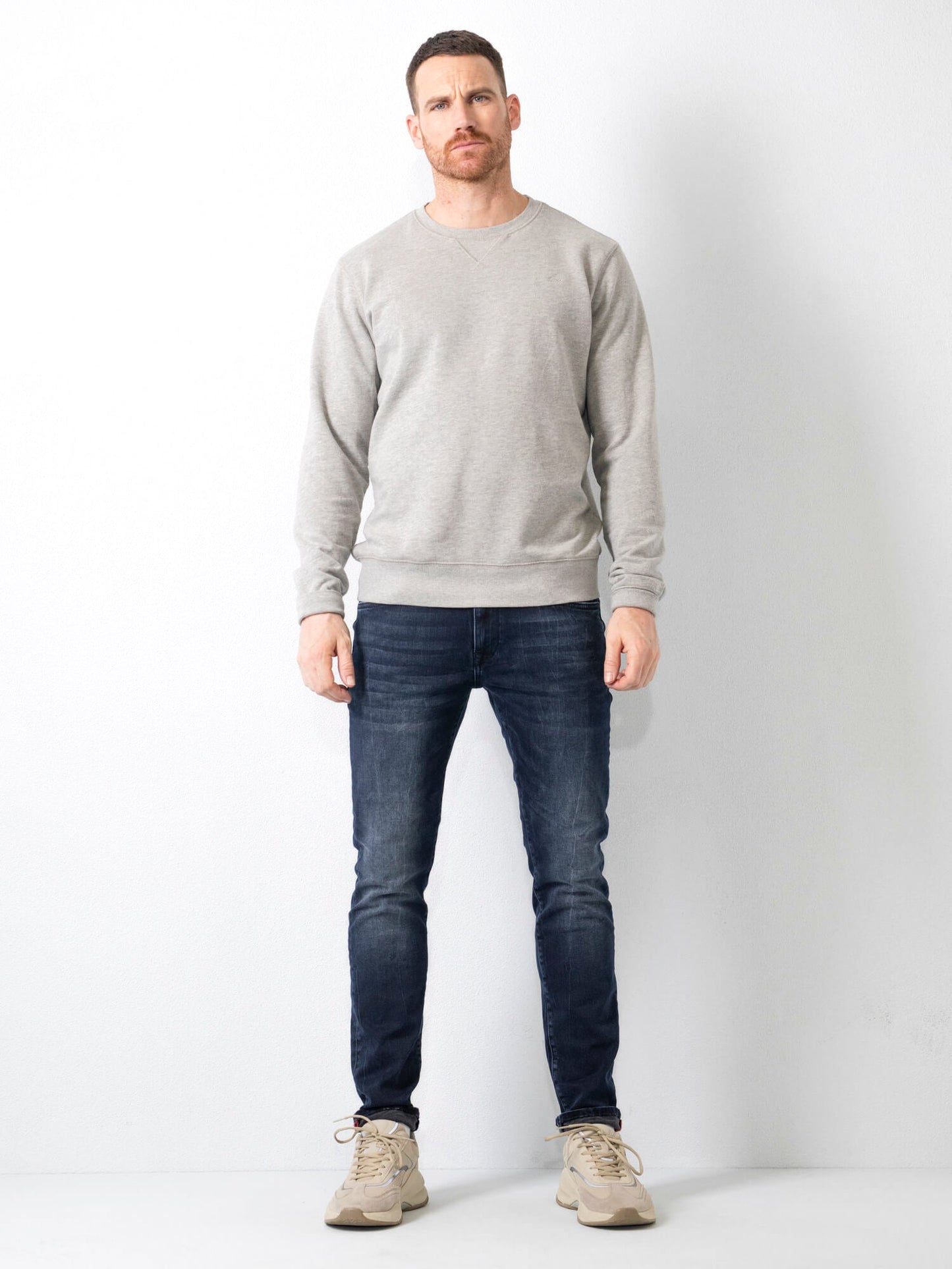 Uomo in jeans scuri e felpa grigia, posa contro un muro bianco. Indossa scarpe sportive beige. Moda casual maschile, abbigliamento autunnale.