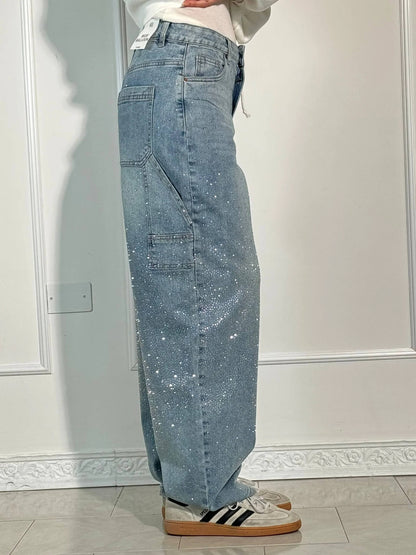 Jeans larghi in denim blu con dettagli glitterati, tasca laterale, abbinati a sneakers bianche e nere. Moda casual, stile urbano.