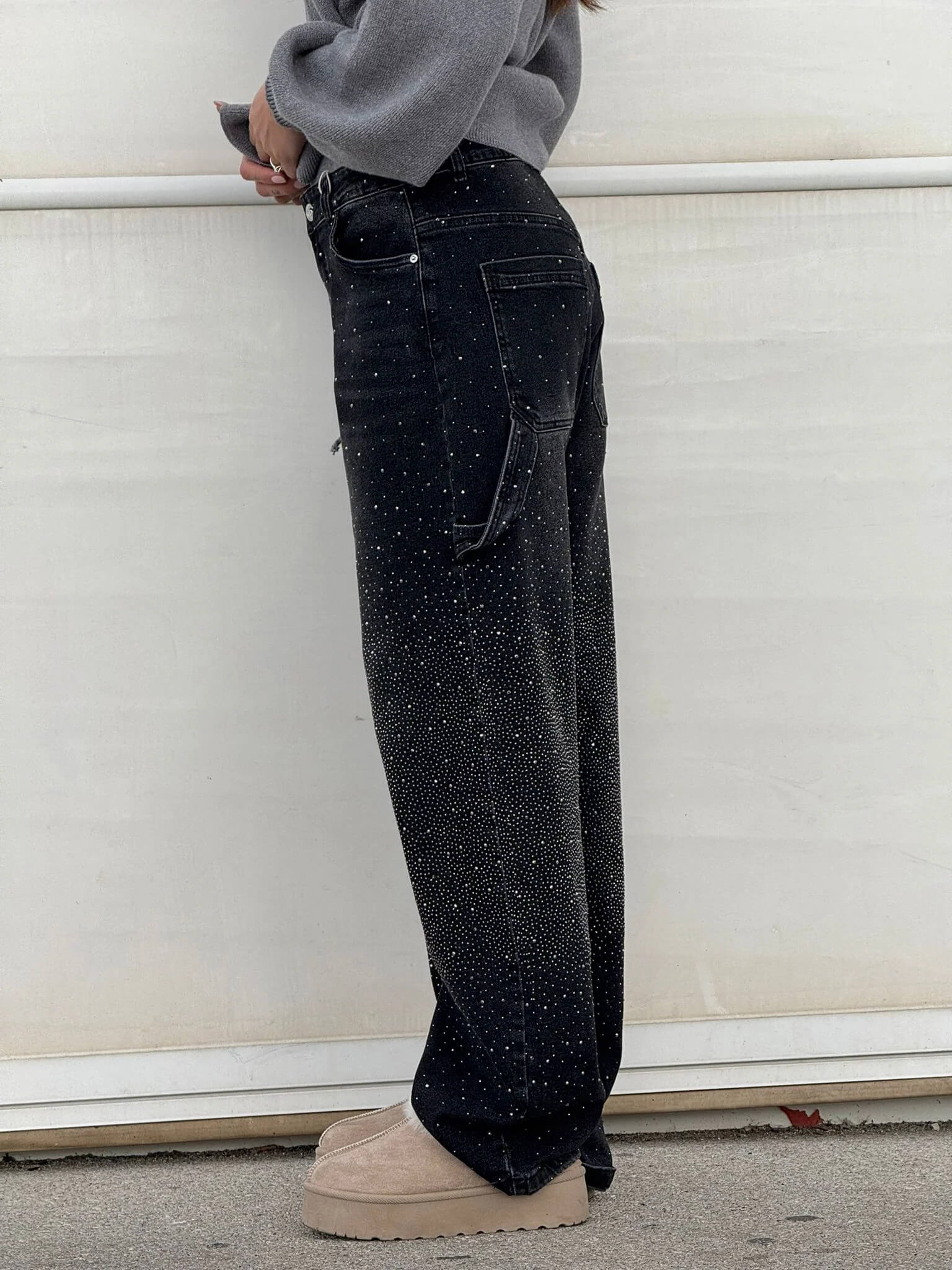 Jeans neri a vita alta con dettagli scintillanti, indossati con maglione grigio e scarpe beige su sfondo bianco. Moda casual elegante.