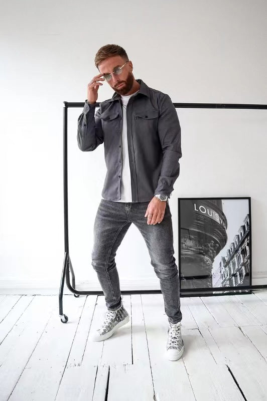 Uomo con camicia grigia e jeans, posa in studio moderno. Occhiali da sole, scarpe da ginnastica bianche. Poster "Louis" sullo sfondo. Moda casual.