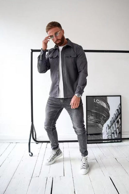 Uomo con camicia grigia e jeans, posa in studio moderno. Occhiali da sole, scarpe da ginnastica bianche. Poster "Louis" sullo sfondo. Moda casual.