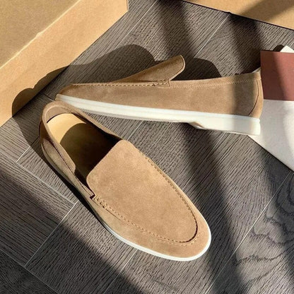Mocassini beige in pelle scamosciata su pavimento in legno, illuminati dalla luce solare. Scarpe eleganti da uomo, stile casual, suola bianca.