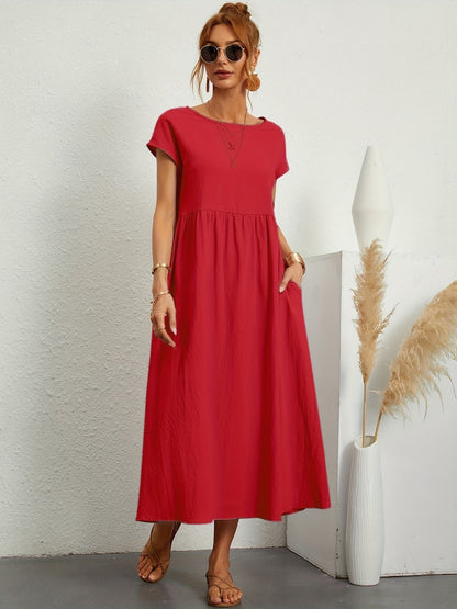 Donna con abito lungo rosso, occhiali da sole, sandali, accanto a vaso bianco con pampas. Moda estiva, abbigliamento casual elegante.