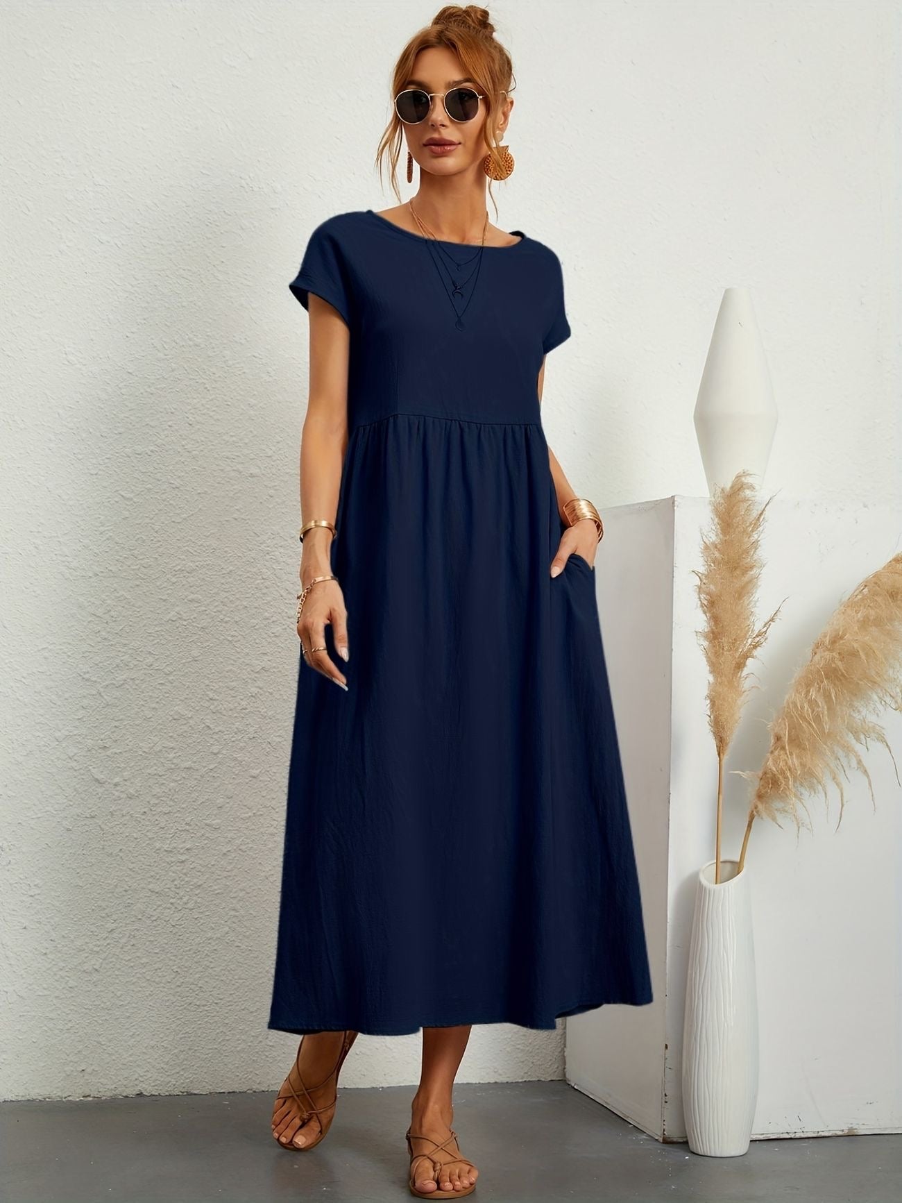 Donna con abito lungo blu navy, occhiali da sole, sandali beige, accanto a vaso bianco con pampas. Moda estiva elegante e casual.
