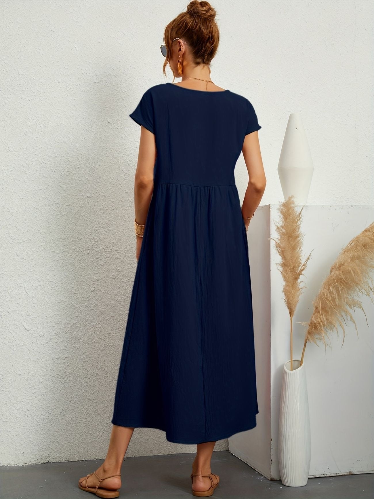 Donna di spalle con abito lungo blu scuro, sandali beige, accanto a vaso bianco con pampas. Moda estiva, abbigliamento casual elegante.