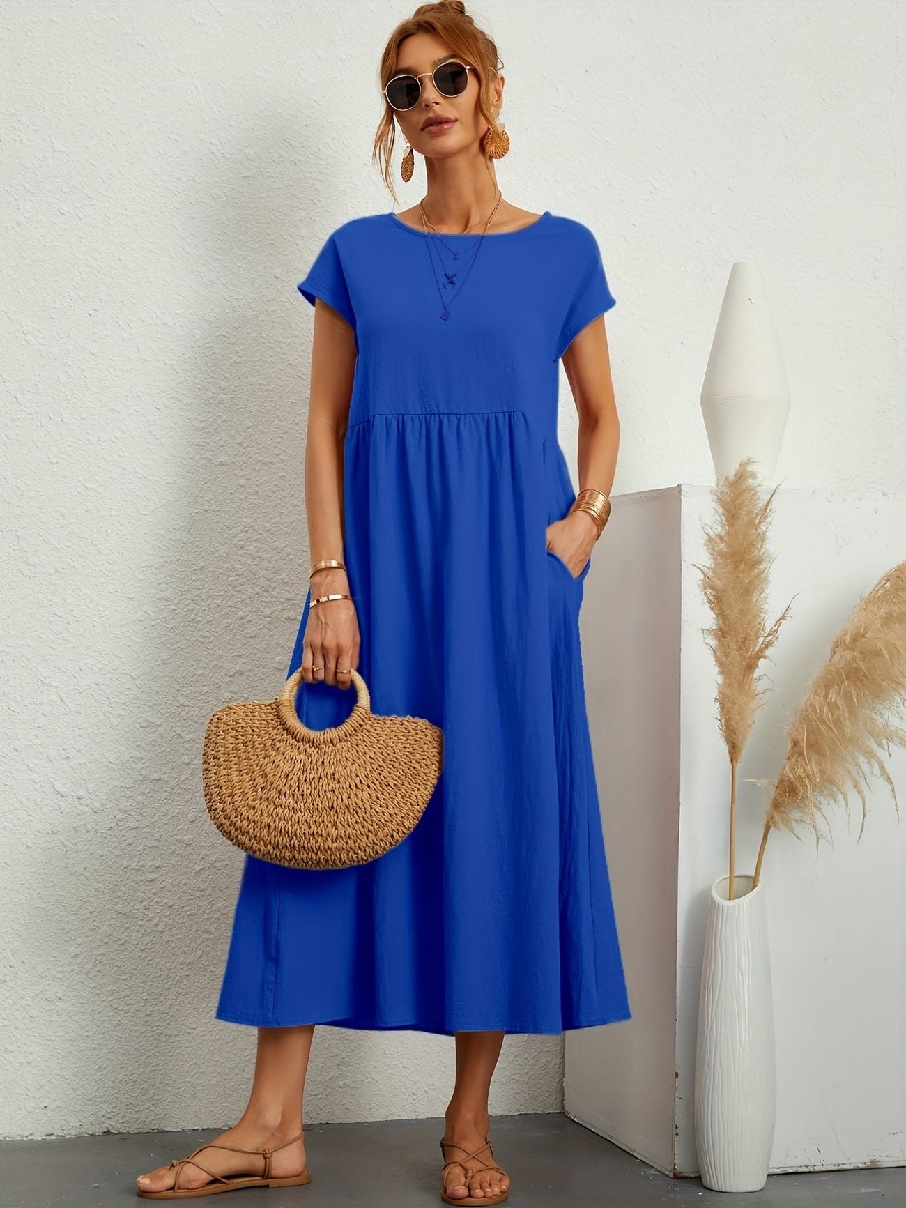 Donna con abito blu lungo, borsa di paglia, occhiali da sole, sandali beige. Sfondo bianco con decorazioni minimaliste. Moda estiva elegante.