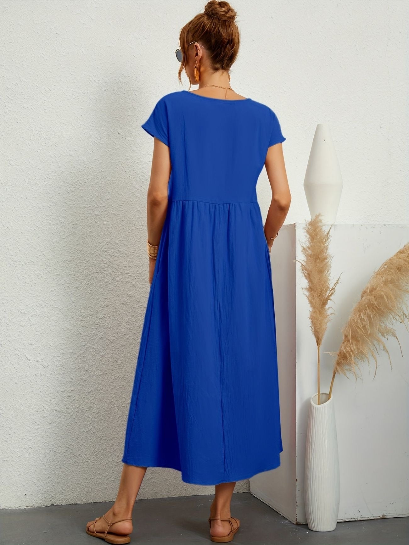 Donna di spalle con abito lungo blu, sandali beige, accanto a vaso bianco con pampas. Moda estiva, abbigliamento casual elegante.
