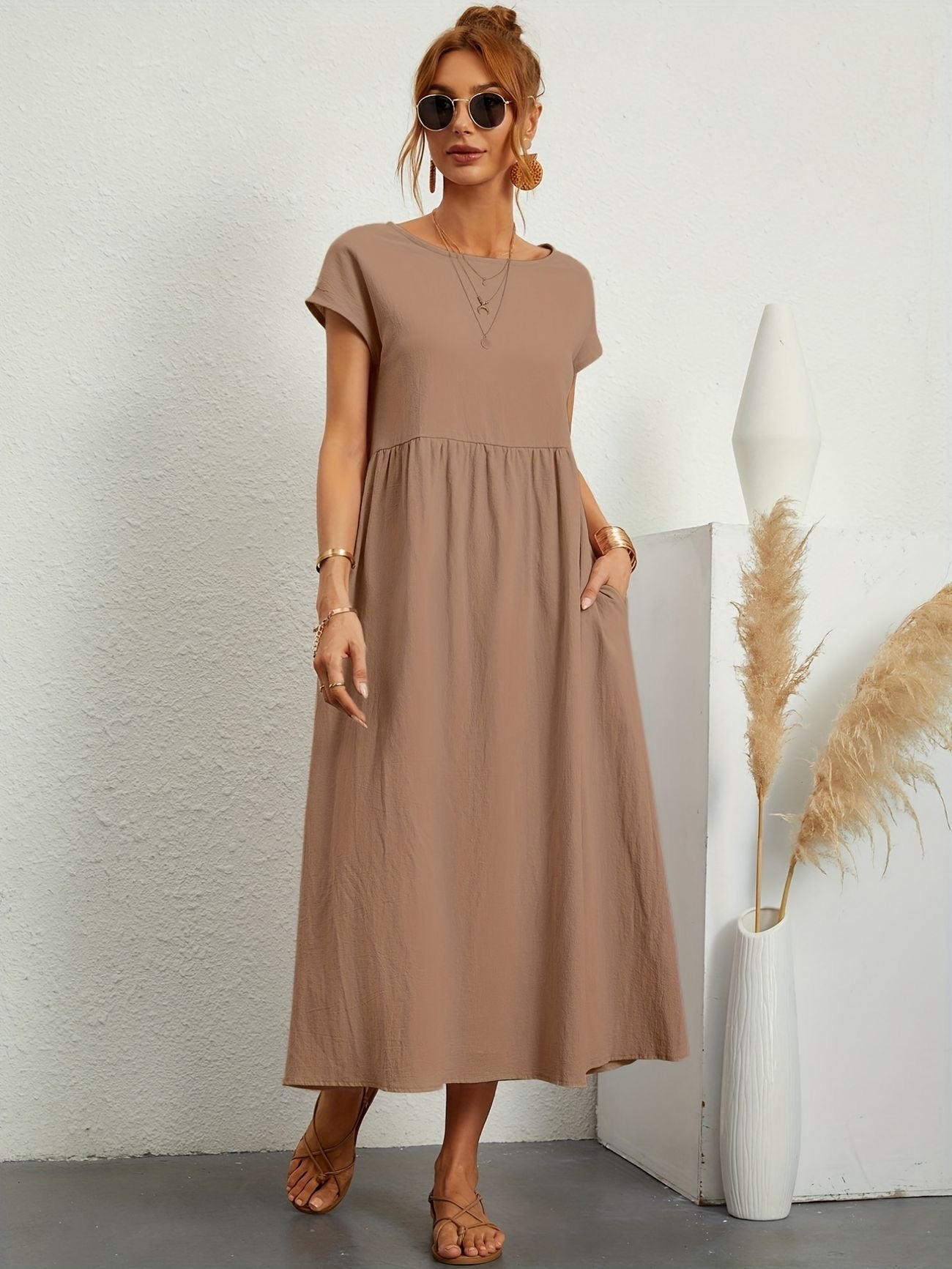Donna con abito lungo marrone, occhiali da sole, sandali, accanto a vaso bianco con pampas. Moda estiva casual, stile minimalista.