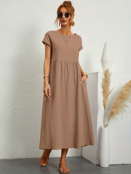 Donna con abito lungo marrone, occhiali da sole, sandali, accanto a vaso bianco con pampas. Moda estiva casual, stile minimalista.