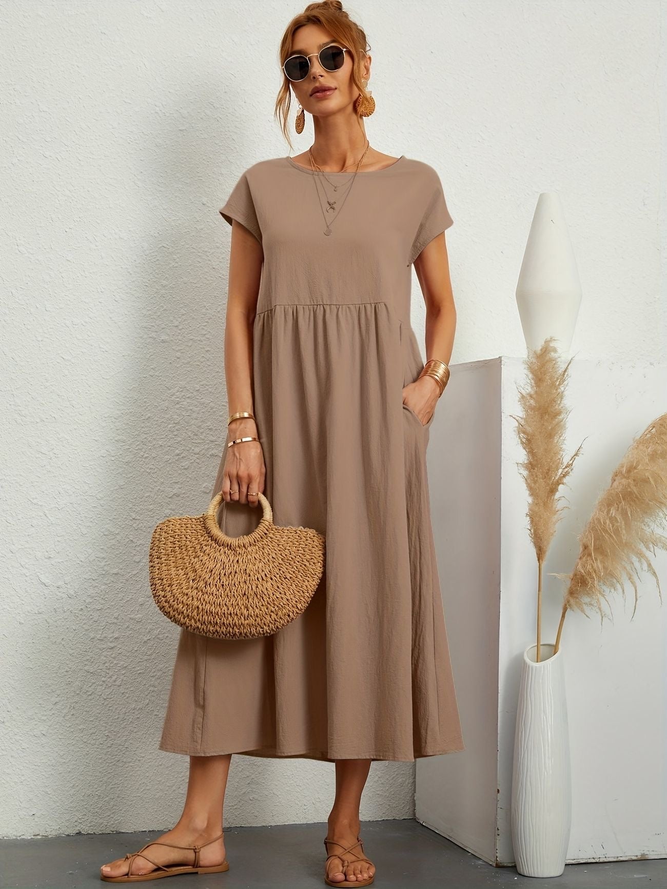 Donna con abito lungo beige, occhiali da sole, borsa di paglia, sandali, accanto a vaso bianco con pampas. Moda estiva elegante e casual.