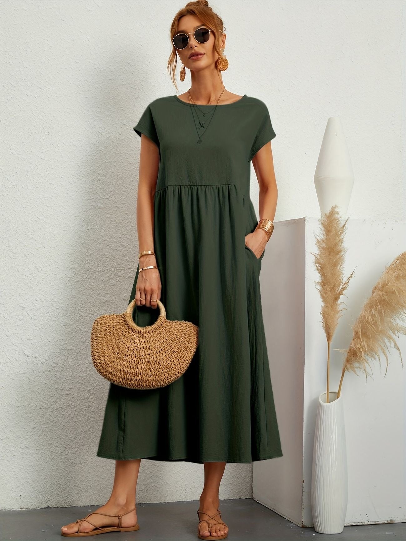 Donna con abito verde lungo, borsa di paglia e sandali, posa accanto a vaso decorativo. Moda estiva, stile casual chic, accessori eleganti.