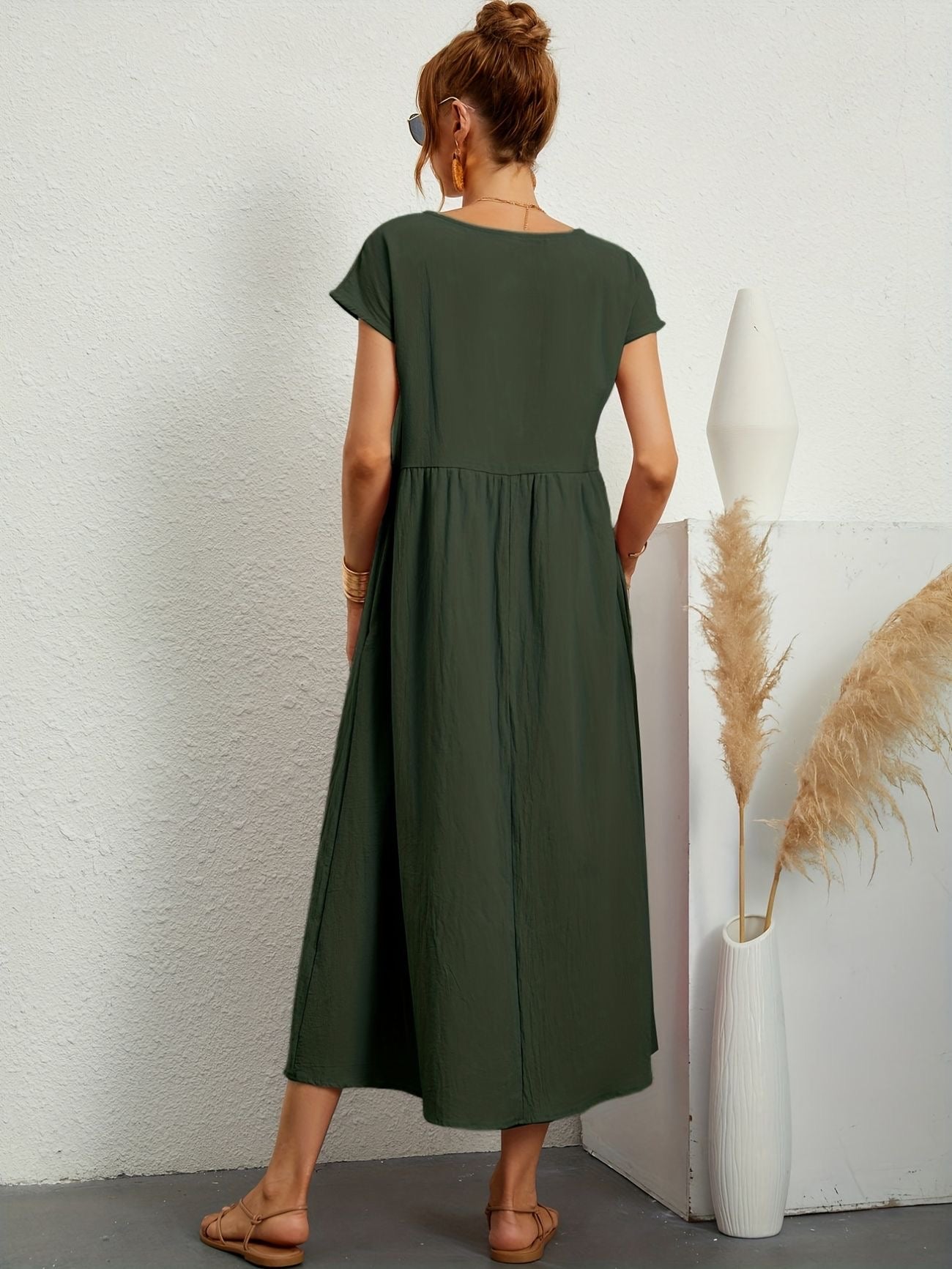 Donna di spalle con abito lungo verde, sandali beige, accanto a vaso bianco con pampas. Stile estivo, moda casual, abbigliamento femminile elegante.