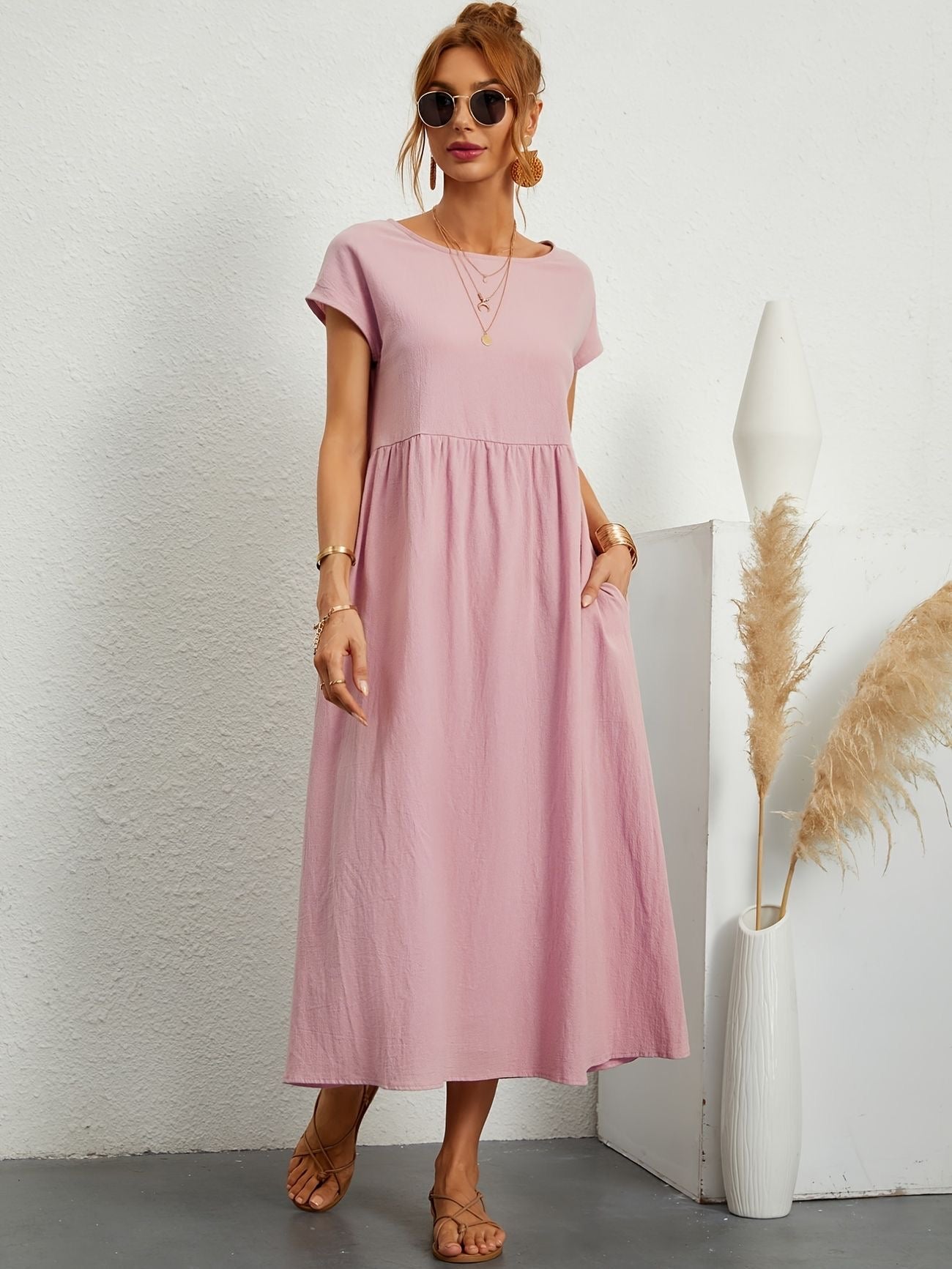 Donna con abito lungo rosa, occhiali da sole e sandali, accanto a vaso bianco con pampas. Moda estiva elegante, stile casual chic.