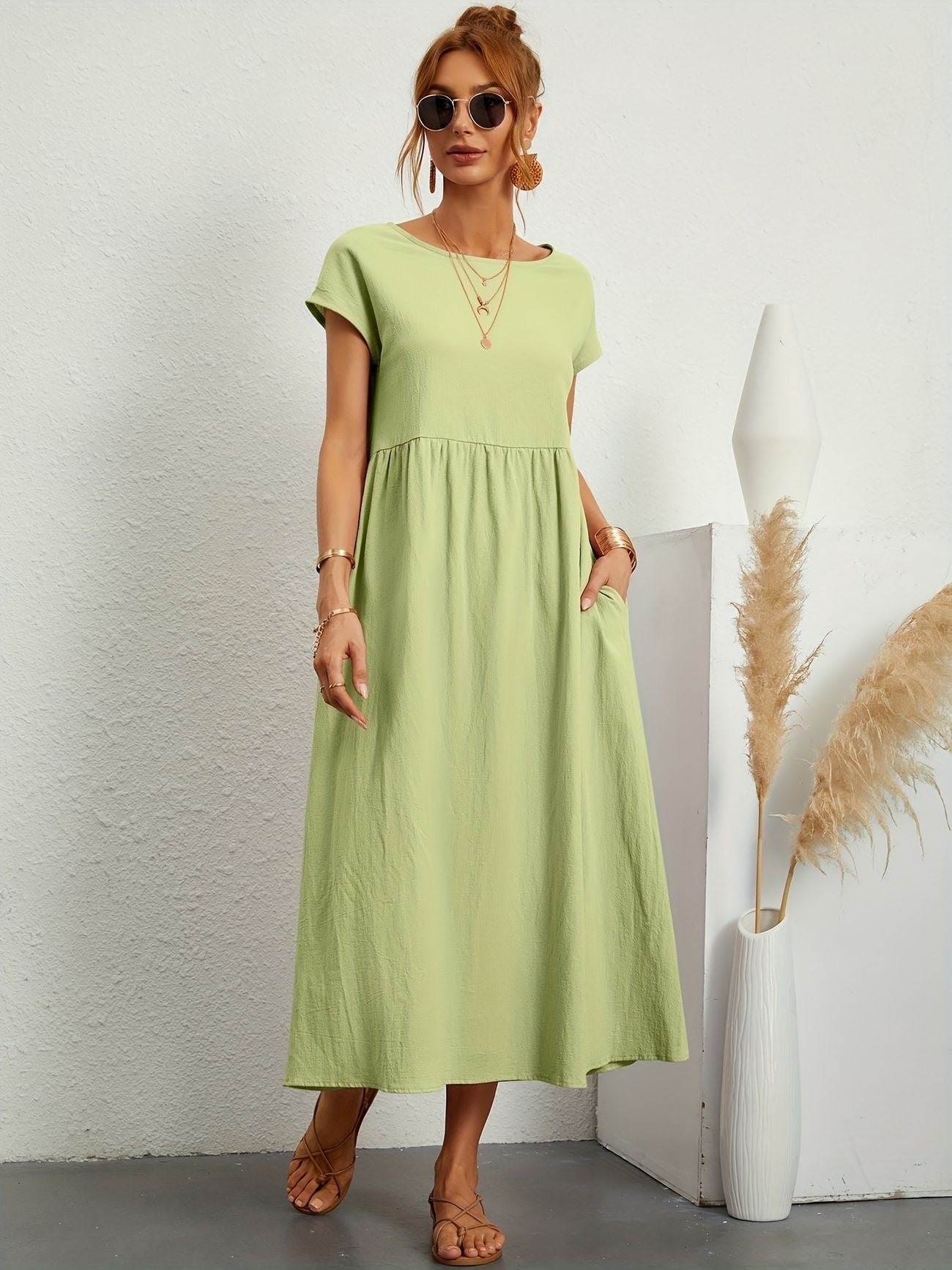 Donna con abito lungo verde chiaro, occhiali da sole e sandali, accanto a vaso bianco con pampas. Moda estiva, stile casual elegante.