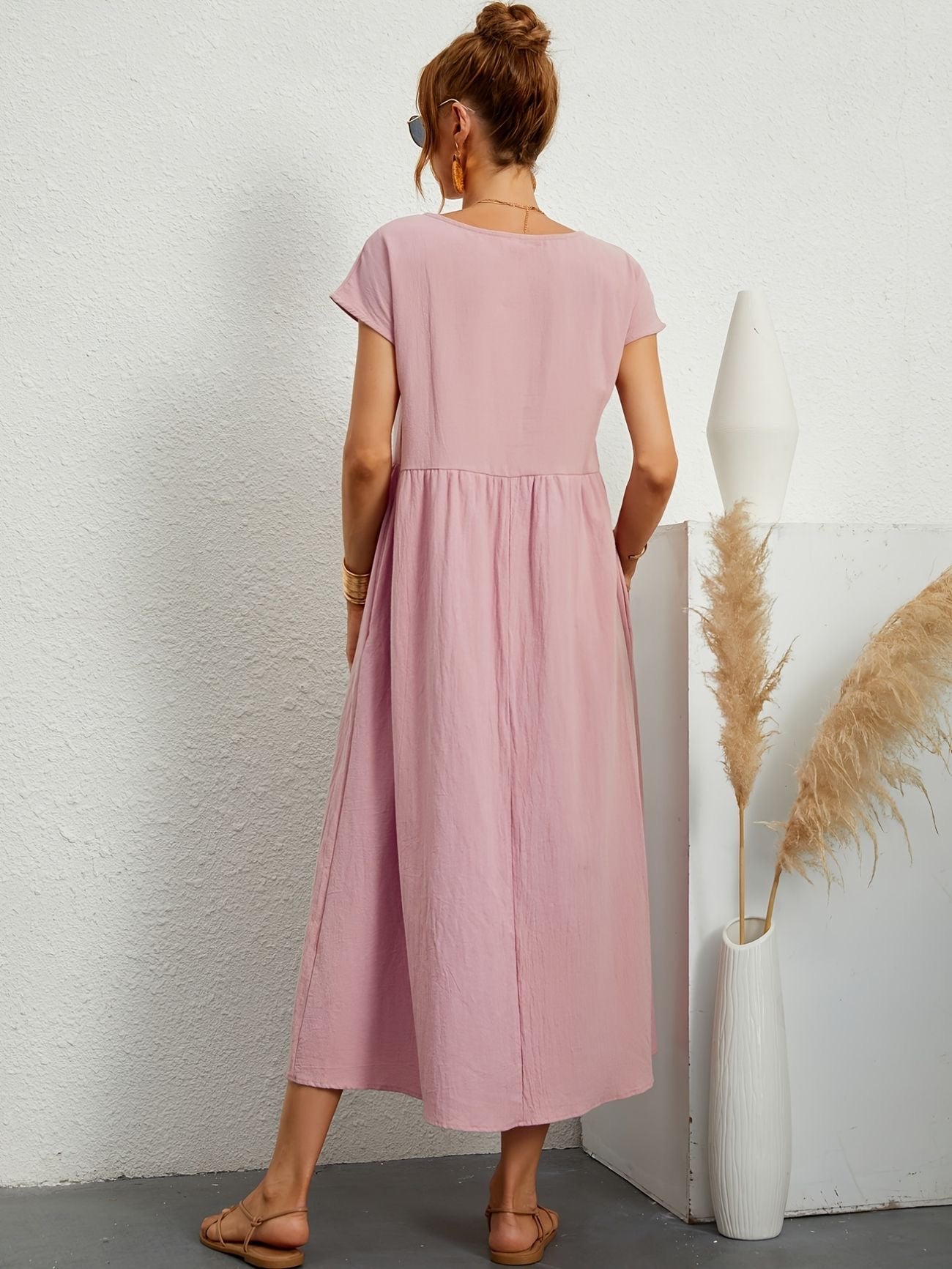 Donna con abito lungo rosa, sandali beige, accanto a vaso bianco con pampas. Moda estiva, abbigliamento casual, stile minimalista.